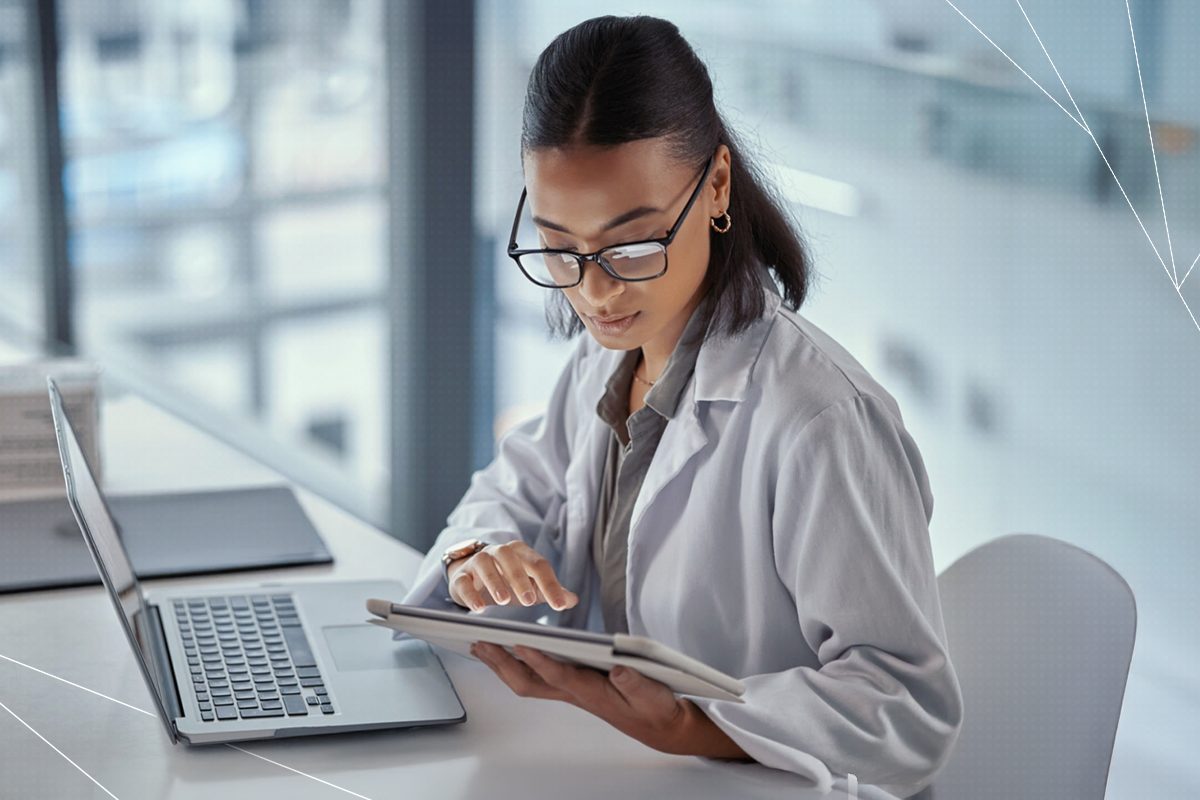 Mulher negra sentada em uma cadeira de escritório segura um tablet com uma mão enquanto a outra digita na tela, com ambos os braços apoiados em uma mesa com notebook, trazendo o contexto de análise de riscos na saúde.