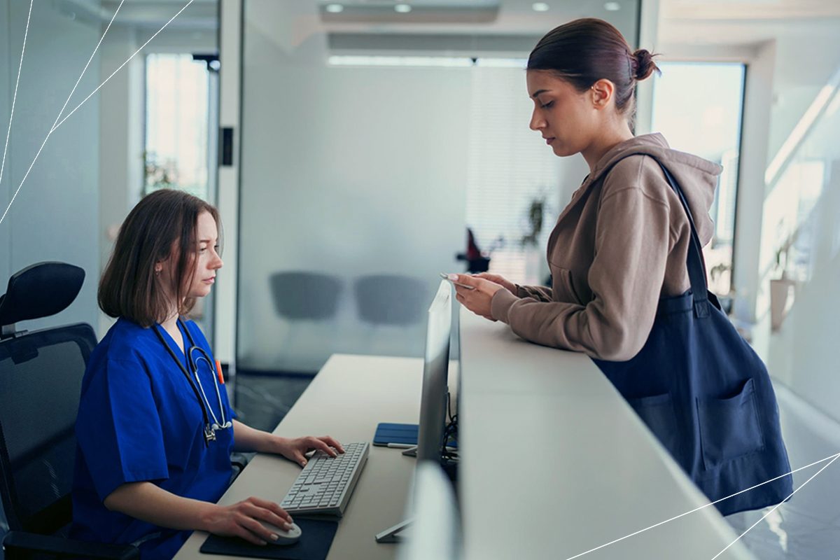 Uma mulher atende uma paciente enquanto olha para o monitor de um computador, digita no teclado e segura o mouse, trazendo o contexto de automação na saúde.