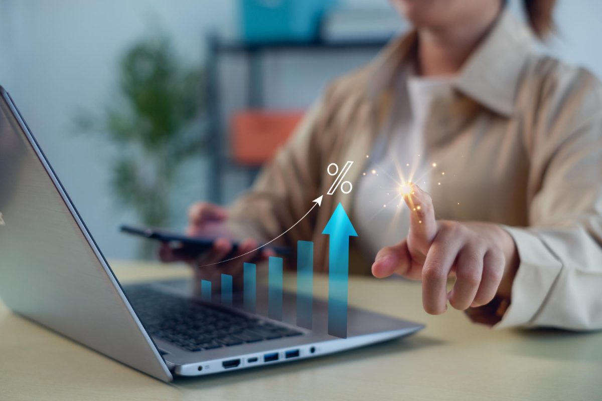 Um dedo da mão de uma pessoa saindo faísca enquanto a outra mão segura um celular, com um notebook sobre a mesa aberto apresentando um gráfico de crescimento à sua frente, trazendo o contexto de crescimento do crédito apontado pelo INDC da Neurotech.