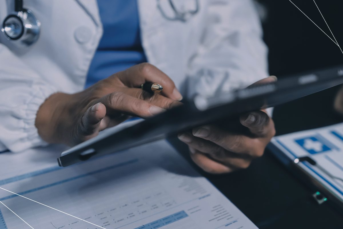 Um médico segura um tablet com uma mão e uma caneta com a outra, enquanto apoia ambas sobre uma mesa de consultório repleta de documentos, trazendo o contexto de sinistralidade em planos de saúde.